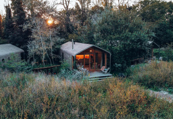 Tiny house immersa nella natura tra erba alta e alberi, con veranda illuminata e tramonto sullo sfondo.