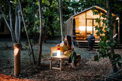 Vrouw op een bankje bij een tiny house in het avondbos, omgeven door bomen en gezellige verlichting.