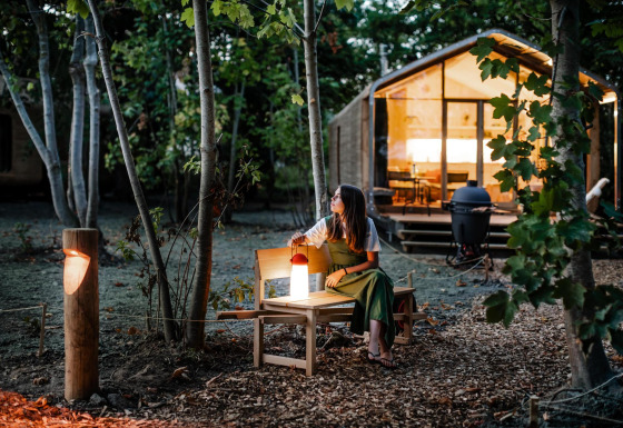 Mujer sentada en un banco junto a una tiny house en el bosque al atardecer, rodeada de árboles y luces.