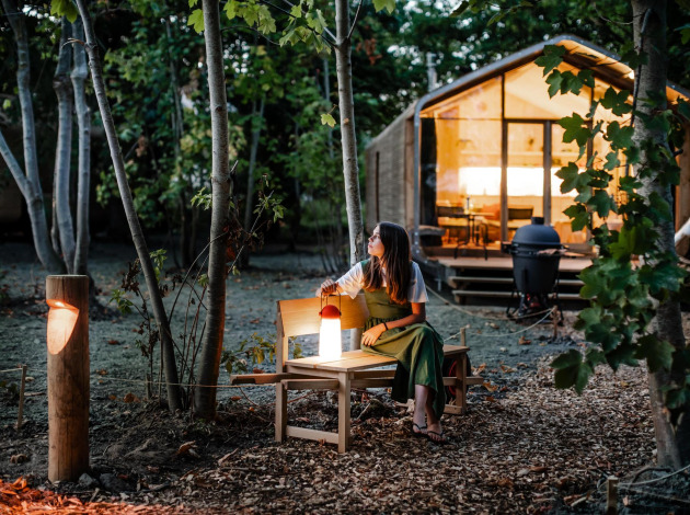 Vrouw zit op een bankje bij een tiny house in het avondbos, omringd door bomen en warme verlichting.