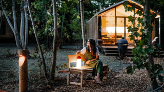 Mujer sentada en un banco junto a una tiny house en el bosque al atardecer, rodeada de árboles y luces.