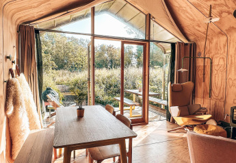 Gezellig, licht interieur van een tiny house met grote ramen, eettafel, zetel en houtkachel.