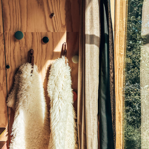 Zonnestralen in het tiny house, met wollen jassen aan haken en gordijnen bij het raam.