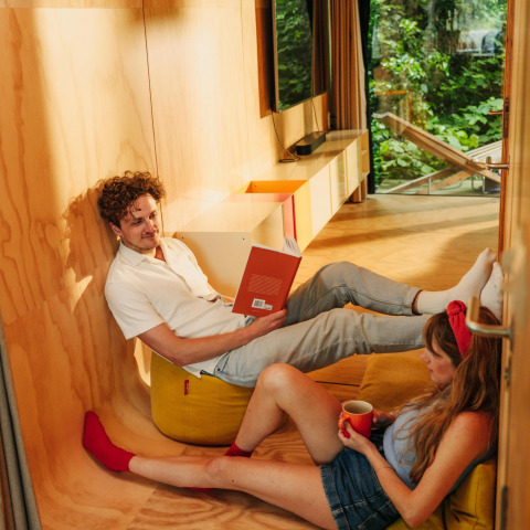 Una pareja se relaja en una pequeña casa de madera; uno lee un libro y el otro disfruta de una taza de café.
