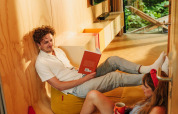 A couple relaxes in a cozy tiny house with wooden walls; one reads a book while the other enjoys coffee.