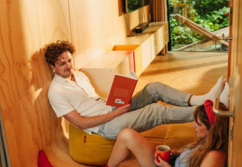 Una coppia si rilassa in una mini casa in legno; uno legge un libro e l’altro gode un caffè.