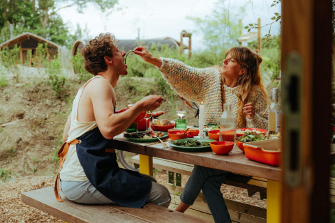 Twee personen genieten buiten van eten aan een picknicktafel bij tiny houses, de vrouw voedt de man.