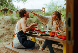 Deux personnes partagent un repas en plein air à une table devant des tiny houses, la femme nourrit l'homme.