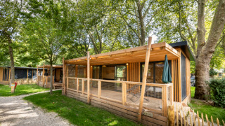 Mobile home Columbia met houten terras, omgeven door hoge bomen en gras in een groene en zonnige omgeving.