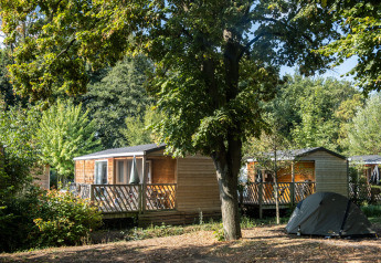 Mobilhjem Cottage omgivet af træer og grønne planter med telt opslået i forgrunden i en rolig natur.