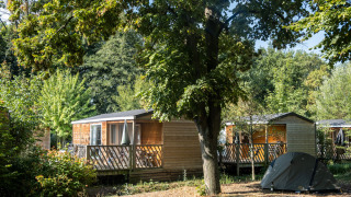 Mobile home Cottage omgeven door bomen en planten, met een tent vooraan in een rustige omgeving.
