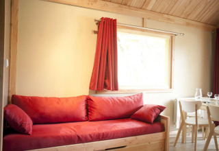Interior de madera clara con sofá y cortinas rojas en el Chalet Montana, Camping de l'Ill - Colmar, Francia.