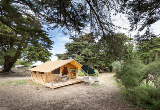La tienda safari Tente Toile&Bois Classic se encuentra instalada en un camping rodeado de árboles altos.