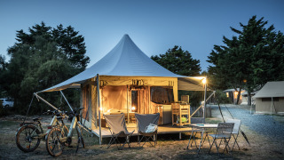 Tente safari Panama éclairée avec entrée, vélos et coin salon extérieur dans un camping en soirée.