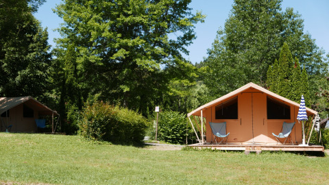 Tente safari Bungalow CLASSIC IV sur un camping verdoyant, entourée d'arbres, avec des chaises à l’extérieur.