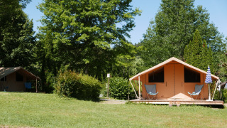 Tenda bungalow safari CLASSIC IV in campeggio erboso, circondata da alberi, con sedie davanti.