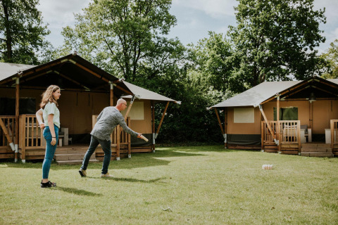 Dos personas juegan a un juego de césped frente a tiendas safari en Camping Si-Es-An, Países Bajos, entre árboles.