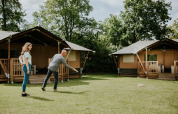 Dos personas juegan a un juego de césped frente a tiendas safari en Camping Si-Es-An, Países Bajos, entre árboles.