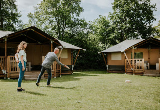 Dos personas juegan a un juego de césped frente a tiendas safari en Camping Si-Es-An, Países Bajos, entre árboles.