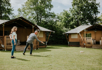 Due persone giocano a un gioco sul prato davanti a tende safari al Camping Si-Es-An nei Paesi Bassi, tra alberi.