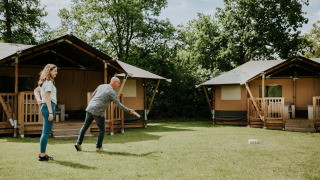 Due persone giocano a un gioco sul prato davanti a tende safari al Camping Si-Es-An nei Paesi Bassi, tra alberi.