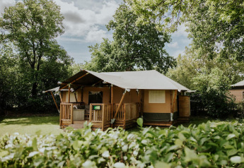 Tienda safari en Camping Si-Es-An, Países Bajos, rodeada de vegetación y árboles bajo un cielo azul.