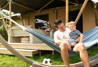 Padre e figlio rilassati sull'amaca davanti a una tenda safari al Camping Si-Es-An nei Paesi Bassi.
