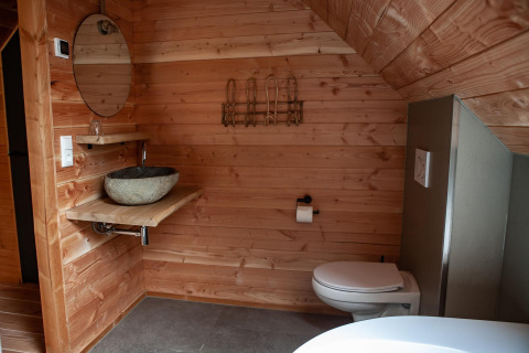 Baño revestido de madera con lavabo de piedra, espejo redondo y inodoro en Heydehuis + Jacuzzi glamping.