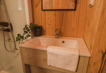 Baño con paneles de madera, lavabo blanco, planta, espejo y ducha en Lodgetent Beaufort, Walsdorf.