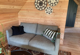 Grå sofa med pyntepuder, vægdekorationer og træpaneler i et hyggeligt sommerhus, Sandberghe, Holland.