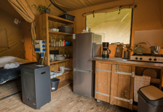 Zona de cocina en tienda safari con armarios de madera, nevera, cocina de gas y cama al fondo.