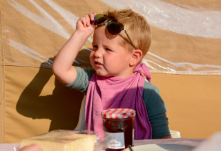 Niño con pañuelo rosa y gafas de sol sentado en mesa dentro de tienda safari en Luxemburgo.