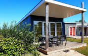 Moderne lodge met ruime, overdekte veranda en tuinstoelen, omgeven door groen en een helderblauwe hemel.