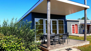 Moderne lodge met ruime, overdekte veranda en tuinstoelen, omgeven door groen en een helderblauwe hemel.