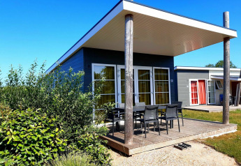 Moderne lodge met ruime, overdekte veranda en tuinstoelen, omgeven door groen en een helderblauwe hemel.