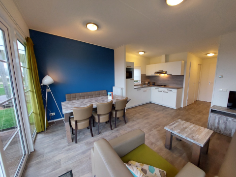 Salon de lodge moderne avec cuisine ouverte, mur bleu, table à manger, fauteuils et grandes fenêtres.