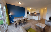Salon de lodge moderne avec cuisine ouverte, mur bleu, table à manger, fauteuils et grandes fenêtres.