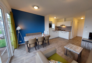 Salon de lodge moderne avec cuisine ouverte, mur bleu, table à manger, fauteuils et grandes fenêtres.