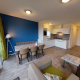 Moderne lodge-woonkamer met open keuken, blauwe accentmuur, eettafel, stoelen en grote ramen.