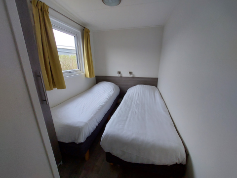 Habitación estrecha en una cabaña con dos camas individuales, sábanas blancas y cortinas amarillas.