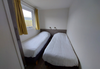 Habitación estrecha en una cabaña con dos camas individuales, sábanas blancas y cortinas amarillas.