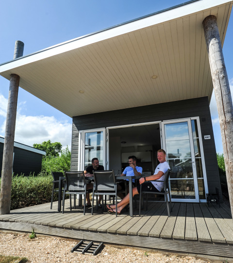 Tre personer slapper af ved et bord på terrassen foran Comfort Lodge på Kompas Camping Westende, Belgien.