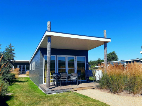 Moderne lodge met overdekte veranda, tuinset en hoge siergrassen onder een heldere blauwe hemel.