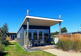 Moderne lodge met overdekte veranda, tuinmeubilair en hoge siergrassen onder een heldere blauwe lucht.