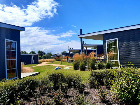 Modern glamping cabins with large windows, green landscaping, and people relaxing under a sunny sky.