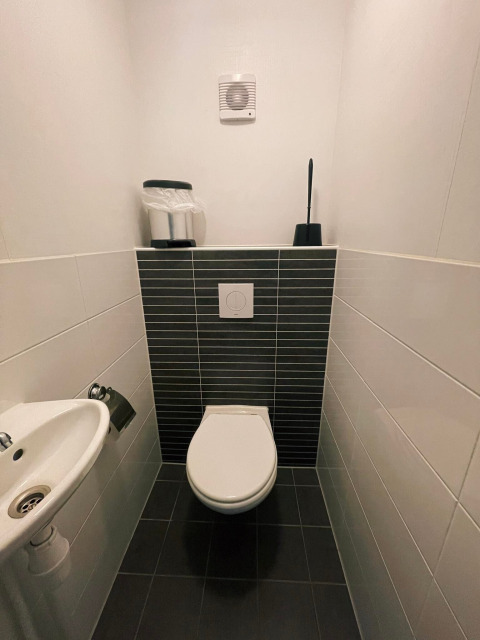 Moderne toiletruimte in Comfort Lodge met zwevend toilet, zwarte tegels en kleine wastafel.