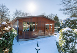 Sneklædt Veluwe lodge i Bungalowpark Het Verscholen Dorp, Holland, omgivet af vintertræer og buske.