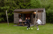 En familie nyder tid udendørs ved en træhytte, mens to børn spiller fodbold ved Camping de Noetselerberg.