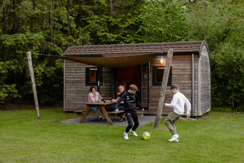 Family relaxing outside a wooden lodge at Camping de Noetselerberg in the Netherlands, kids playing soccer.
