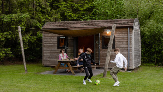 Gezin ontspant bij een houten lodge op Camping de Noetselerberg in Nederland, kinderen spelen voetbal.